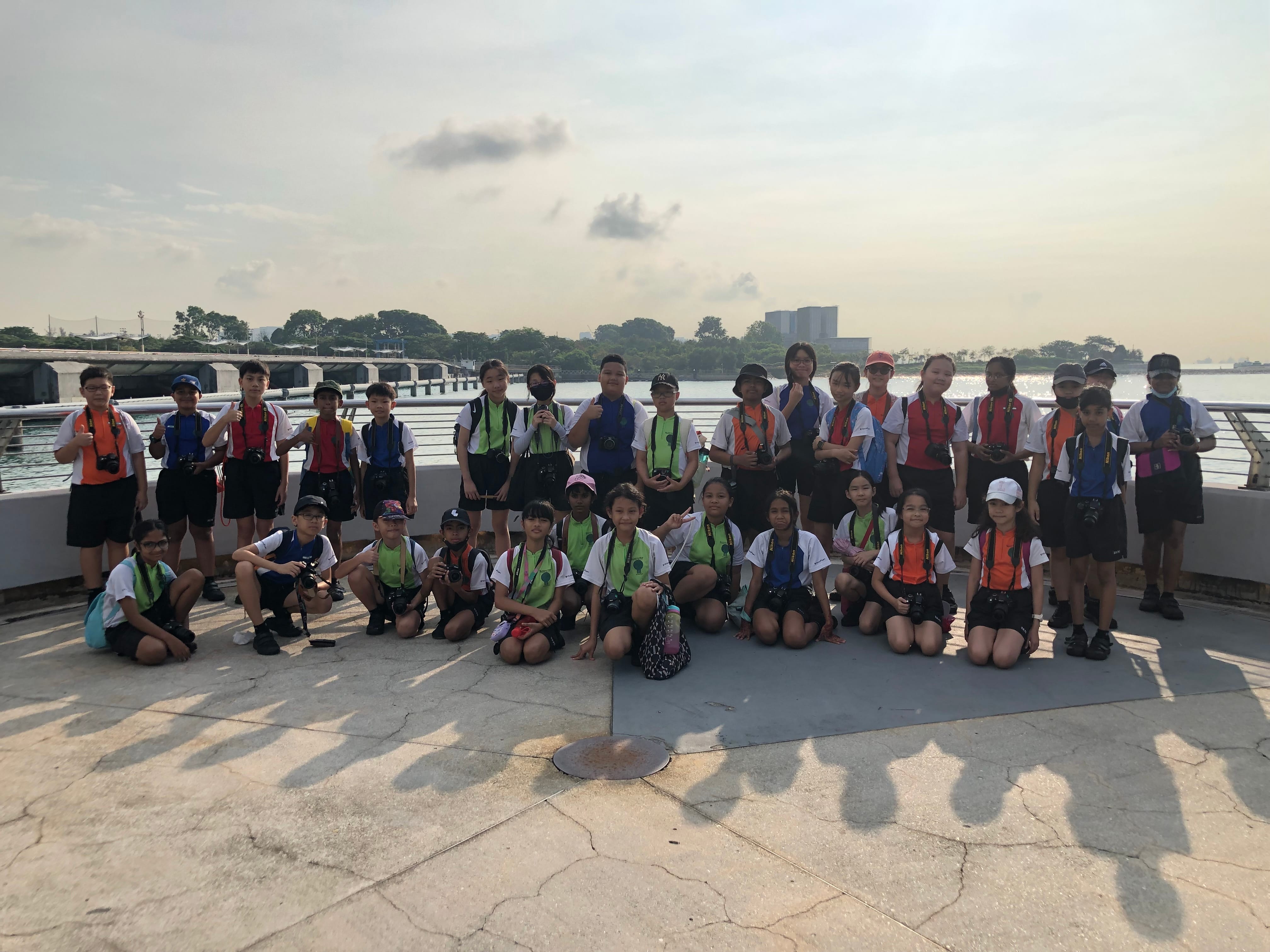 A large group of children with cameras pose on an outdoor waterfront platform with trees and buildings in the background.